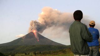 A photo of a volcano erupting in the distance and people watching