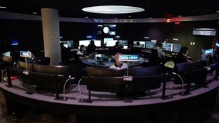 A circular desk arrangement with people sitting on the inside facing outward. Everyone has a double monitor screen and most are wearing a headset. "Nova Control" is written in large silver letters above the workers.