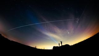 track the iss from Earth, this Photograph shows two silhouettes standing looking up at the sky, the one on the right is pointing up at the white streak of light across the sky. The white streak is the ISS as it flies overhead. Stars can be seen in the background. 