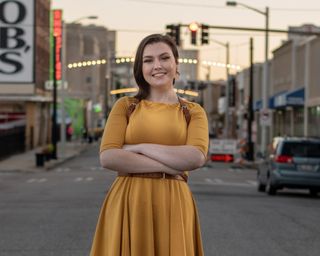 Alyse Stanley, smiling, is standing in an intersection at dusk — with her arms folded across herself.