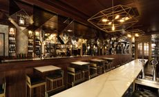 A bar with a long light marble counter, tables, chairs, wall mirrors and wooden walls.