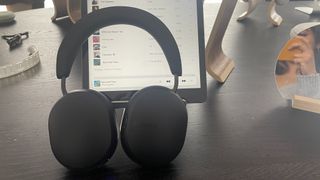 A black pair of Sonos Ace headphones standing on a desk in front of an iPad with a playlist on screen.