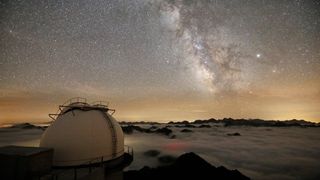 Observatory above the clouds below and the bulge of the milky way stretches across a star-filled sky.