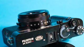 The Fujifilm X100VI mirrorless camera lying on its side against a blue background propped up using the lens cap.