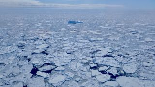 Antarctic sea ice is seen breaking up. A lone iceberg sits in the center of the ocean.