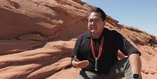 man squatting in front of a complex rock formation with blue sky