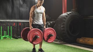 Man holding a trap bar with plates in his hands performing a farmer&#039;s walk along grass in a gym