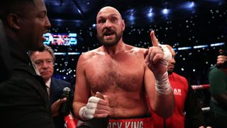 Tyson Fury, wearing red shorts emblazoned with &#039;Gypsy King&#039;, stands in a boxing ring pointing ahead of the Fury vs Usyk live stream 