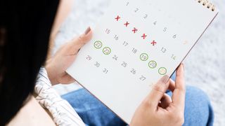 close up on a woman's hands holding a calendar, on which there are five days marked with a red X (representing her last period) and five days marked with green circles (representing ovulation)