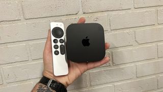 A black Apple TV 4K streaming device and white remote held in one hand in front of a white brick wall.