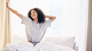 How to get up earlier image shows a woman with curly black hair stretches and smiles as she wakes up early on a sunny summer's morning