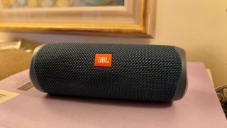 A blue JBL Flip 5 speaker on a purple surface in front of a wall-hung picture frame.