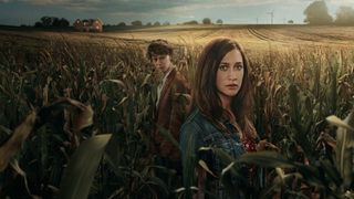 Vera (Julia Ragnarsson) and Isak (Erik Enge) stand in a wheat field with a house and wind turbines in the background in End of Summer