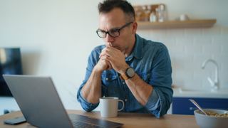 Man sitting at a table and looking at a laptop, holding one hand in the other in front of his face and looking concerned