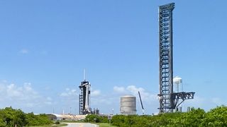 Green shrubbery and sliver of road form a low horizon below a sunny blue sky. In the distance left of center, a short white rocket stands adjacent to a black tower. Much taller, right of center, reaching to the top of the image, a steel tower with black-barred extension arms reaching out near its base.