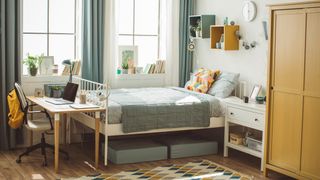 Image shows a brightly coloured dorm room with a clean and comfy mattress placed on a white bedframe and dressed with brightly coloured cushions and a grey throw 