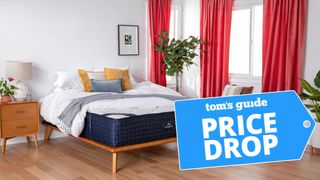 Image shows the DreamCloud memory foam mattress placed on a wooden bedframe in a white bedroom dressed with red curtains, and with a blue price drop sales image overlaid on the bottom right hand corner