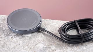 The top of the Anker Wireless Charging Pad, which is sitting on a grey stone-effect surface, against a light pink background. Its cable is neatly wound and secured with a thin black elastic band. 