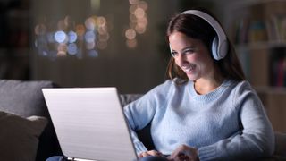 A woman sitting on a coach and on her laptop, wearing headphones, at night time