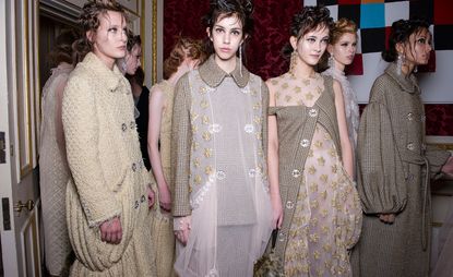 Eight female models wearing ivory, cream and brown embellished and patterned pieces from Simone Rocha&#039;s collection. In the background there is a deep red patterned wall and a white and gold door and frame