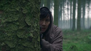 A hooded man peeks out from the tree he is hiding behind. In the background there is a forest full of tall trees.