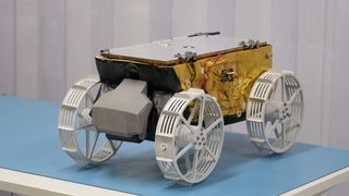 A gold-foiled rover with white wheels sits on a white table covered in a blue tablecloth.