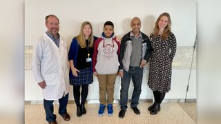 A photo of five people standing in a line: a male doctor in a white lab coat, a woman in professional clothing and a lanyard, a 11-year-old boy, the boy's father, and another woman in professional attire