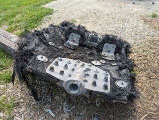 a large black piece of fiberglass covered in metal bolts and plates lies on the ground beside a trail leading into a forest. mountains can be seen rolling in the distance