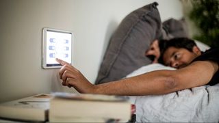 A man reaching out of bed with grey pillows to a bedside smart bed control panel on the wall.