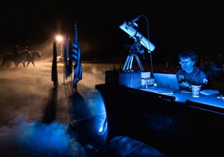 a man sits at a table with a laptop and telescope during a nighttime event. the american flag and two people on horseback are visible to his left.