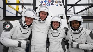 four people in white spacesuits with the visors up pose and smile in front of a white cone-shaped spacecraft