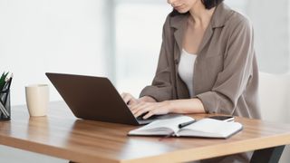 Woman sitting at a desk in a room lit by daylight and working on a computer, her face obscured