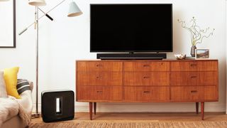 A black Sonos Sub on the floor next to a wooden TV cabinet with drawers. The Sub stands in front of a floorstanding lamp, behind a rug and next to a sofa with a yellow cushion.