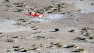 a small black capsule sits in brown desert scrubland, with a scrunched-up orange-and-white parachute on the ground nearby.