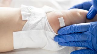 close up on a babies leg as a gloved medical provider places a bandaid on it, as if after a vaccination