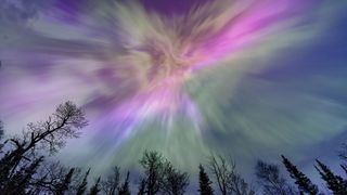 Northern lights appear over a lake in Minnesota in a dark sky overhead shining rainbow of Aurora light with trees below. 