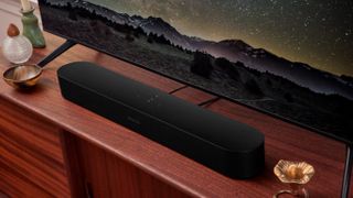 A black Sonos Beam (Gen 2) soundbar on a dark wood TV cabinet in front of a TV showing a nighttime mountain scene.