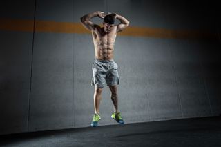 Man performing a burpee against a grey backdrop jumping into the air with his hands behind his head