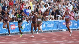 Five women sprint on a track with a large audience in the background