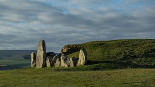 a set of stones line up in a row in the grass. behind is a large hill, far behind are other hills and the cloudy sky in daytime