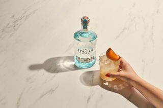 A bottle of Volcan de mi Tierra tequila on a light coloured marble surface. Next to the bottle is a hand holding a glass of a tequila cocktail with ice and a piece of fruit in it