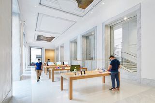 A white room with rows of wooden tables with computers on them, large windows, a staircase and a high pressed roof.