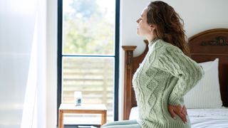 A woman with long dark hair in a light green jumper sits on a soft mattress topper holding her lower back because she has pain there