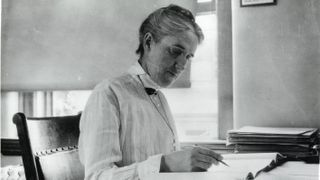 Black and white photograph of Henrietta sitting at a desk wearing a long sleeved blouse, holding a pen in her right hand and looking at some pages on the desk.