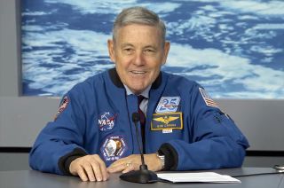 a smiling gray-haired man in a blue jacket sits at a gray table.