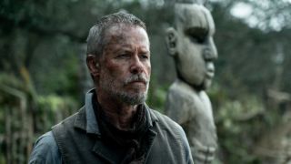 Guy Pearce stands near a outdoor statue with a stoic look on his face in The Convert.