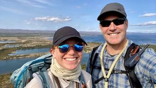 two people in hats and sunglasses smile for a selfie with a lake behind them