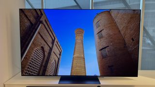 The LG G4 OLED TV on a white stand in a showroom. On the screen is a tower between two large buildings.