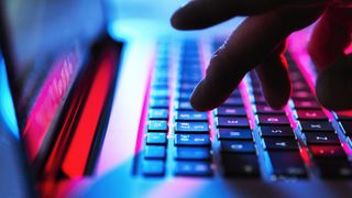 A zoomed-in image of a hand typing on a laptop. The laptop is bathed in red and blue light