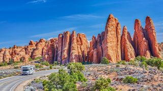 annular solar eclipse road trip. A large orange red rock formations reaching up to the blue sky. A large RV drives along the road in the lower left portion of the image. 